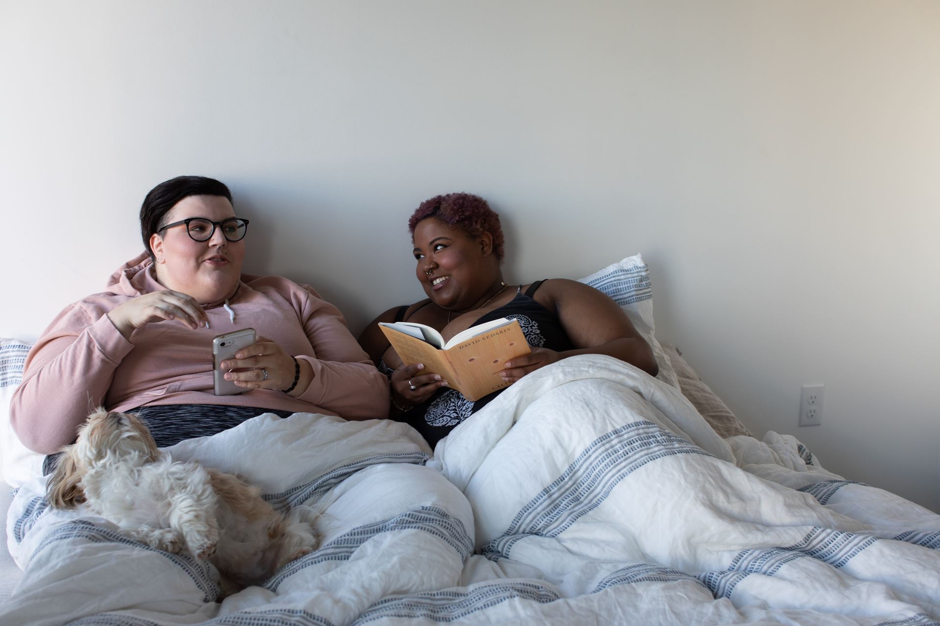 Two larger women sitting up in bed. One is light-skinned, holds a phone, and has a small dog lying in her lap. The other is darker-skinned and holds an open book.