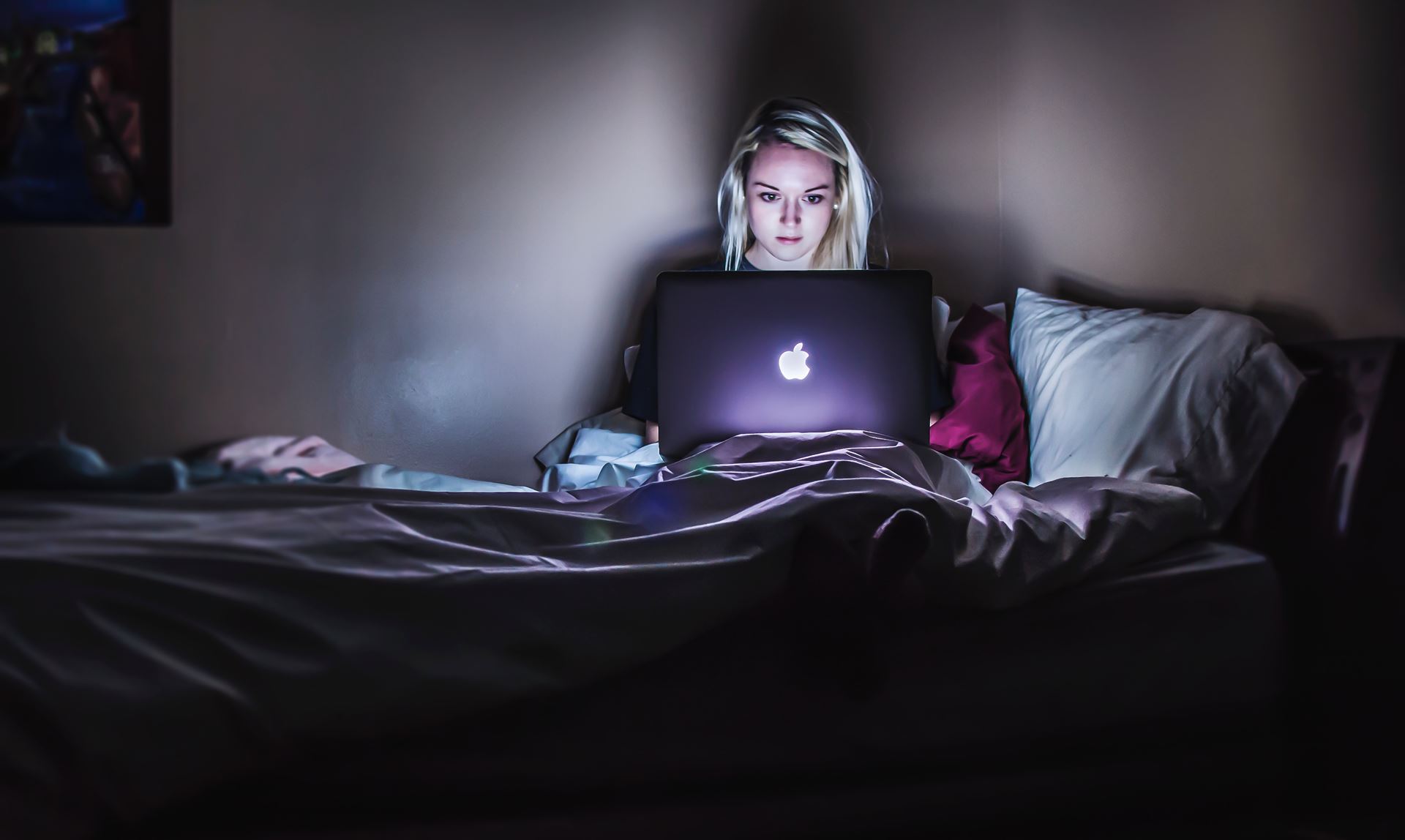 A female-presenting person sitting up in bed on a Macbook laptop. The room is dark, indicating that it is nighttime, with the only light coming from the laptop.