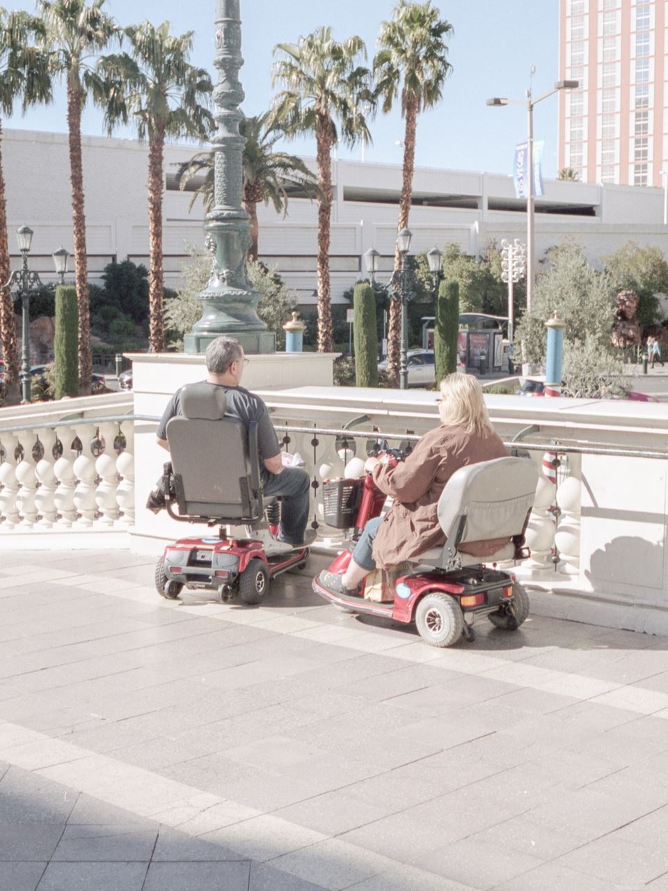 Two older, white people sit in mobility scooters, facing away from the camera and overlooking a street lined with palm trees.