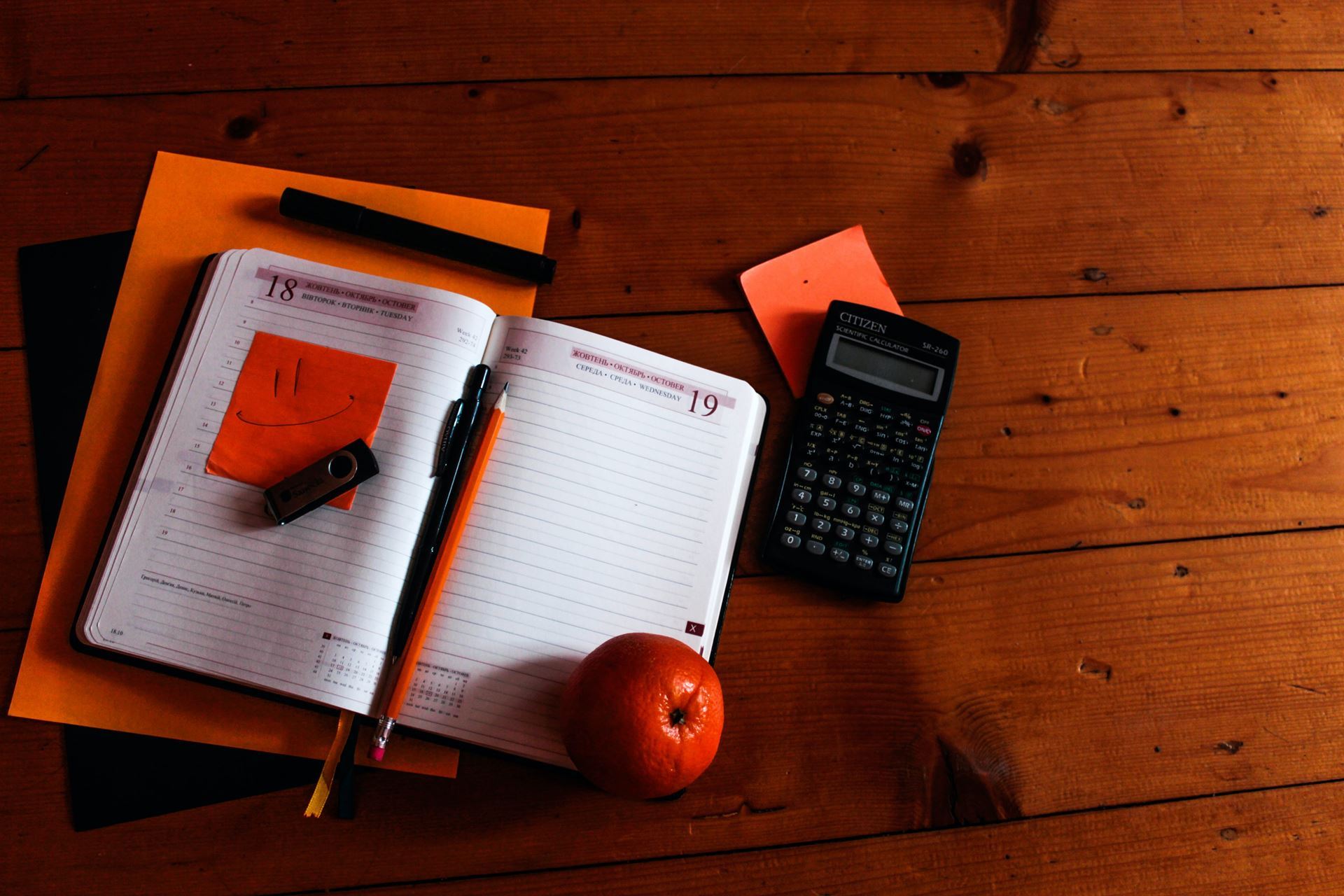 An open notebook, USB stick, pen, pencil, calculator, and apple, all on a dark wooden surface