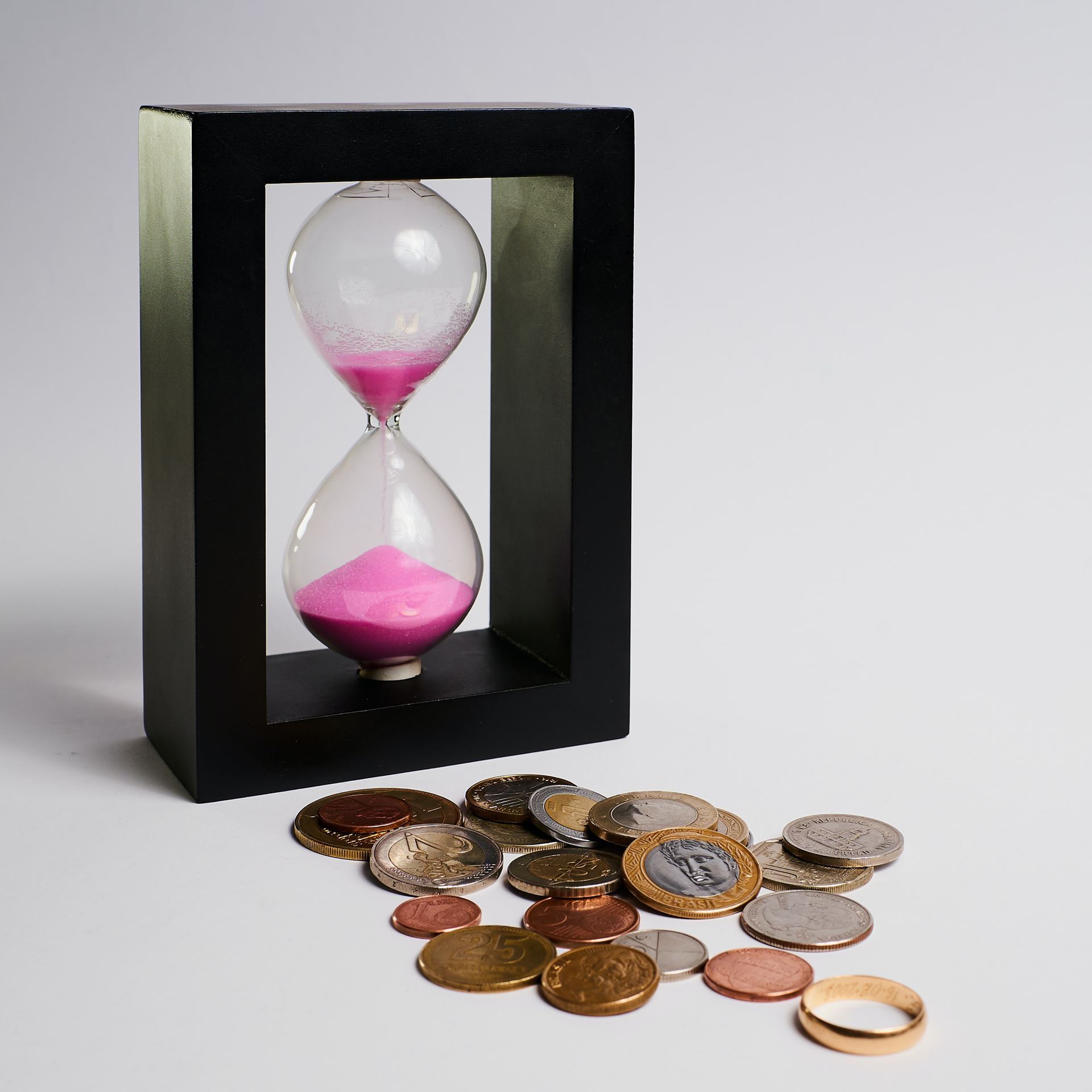 An hourglass with pink sand in it inside a thick black frame, euro coins of various denominations, and a gold ring, all against a light grey background